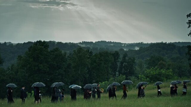 Grieving people in black walking in a field during a burial ceremony on a rainy, overcast day