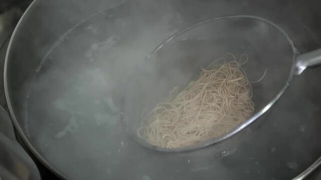 A detailed shot showing the process of cooking traditional thin Asian noodles or vermicelli. The noodles are held in a mesh strainer submerged in boiling water, with visible steam and bubbles capturin