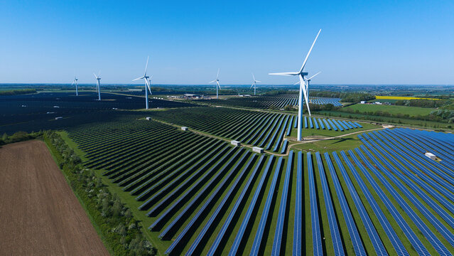 Aerial view of Chelveston Wind Farm featuring rows of solar panels and tall wind turbines under a clear blue sky in Wellingborough, England, United Kingdom.