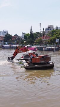 Vertical screen River Excavator Dredging Can Tho Waterfront, Closeup Of Orange Excavator On Floating Pontoon Removing Floating Vegetation And Sediment, Muddy Brown Water, Urban Riverbank