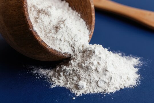 Fine white powder spills from wooden bowl onto dark blue surface. This substance is likely magnesium l-threonate used in brain health research and supplements.
