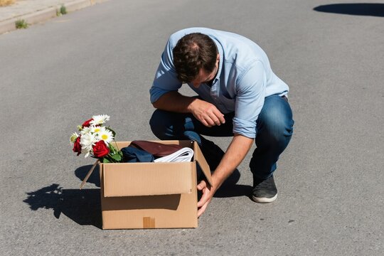 Man with box of personal items on street, sad face, sitting on asphalt. Unemployment situation, career change concept, new beginnings after job loss. Hopeful outlook.