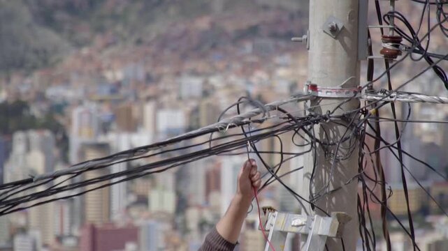 A man's hand makes a makeshift connection to a power cable. The power cable is located on a utility pole alongside a tangled mess of other wires attached to the same post. 