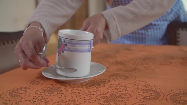 A lady's hands arrange a cup, a saucer, and a teaspoon on a dining table.