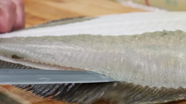 Close-up footage of a skilled chef's hands using a sharp knife to carefully fillet a fresh halibut on a wooden cutting board. This professional technique, often used for high-quality sashimi or sushi 