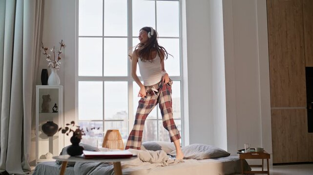 Joyful woman jumping bed home. Happy brunette pyjama having fun listening music