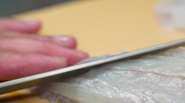 High-angle, close-up shot of a skilled chef's hand using a sharp Japanese knife to delicately slice translucent white fish fillets on a wooden cutting board. The footage showcases traditional Japanese