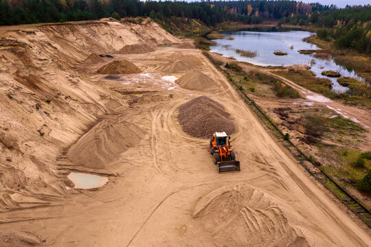 Aerial view of a sand quarry with an orange front loader working near piles of gravel and a calm lake surrounded by autumn forest in Nemencine, Vilnius County, Lithuania.