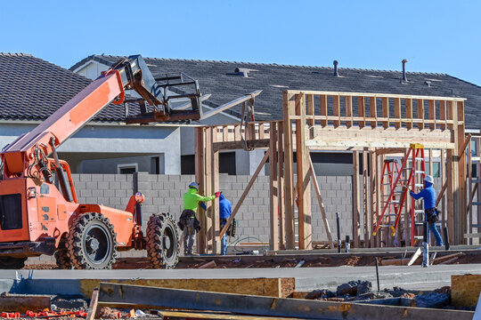 Telescopic forklift lifting framing materials while workers assemble wood framing at a residential construction site, representing structural assembly, material handling, and housing development