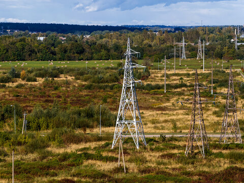 Aerial view of high-voltage power lines and electricity pylons crossing a rural landscape with green fields and a dense forest background in Nemencine, Vilnius County, Lithuania.