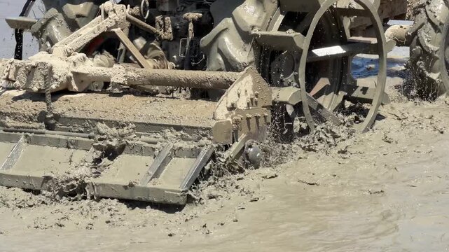 Close view of tractor blades stirring mud and water while shaping the paddy surface for planting. Agricultural machinery, rice farming.