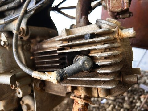 Detailed close-up of an old motorcycle engine showing a spark plug connection, cooling fins, and a heavily rusted exhaust pipe.