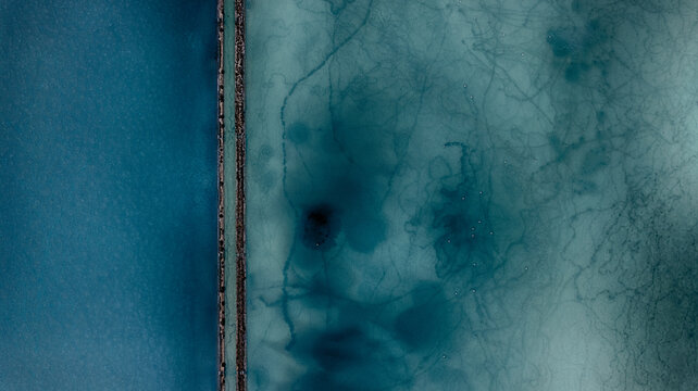 Aerial view of the salt pans with vibrant turquoise water and organic sediment patterns divided by a narrow stone wall in Cagliari, Sardinia, Italy.