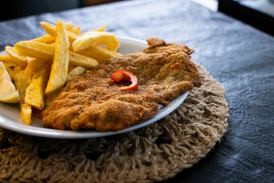 Detalle macro de una milanesa crujiente con enfoque selectivo en la textura del empanado, acompa&ntilde;ada de papas fritas y una rodaja de lim&oacute;n fresco.