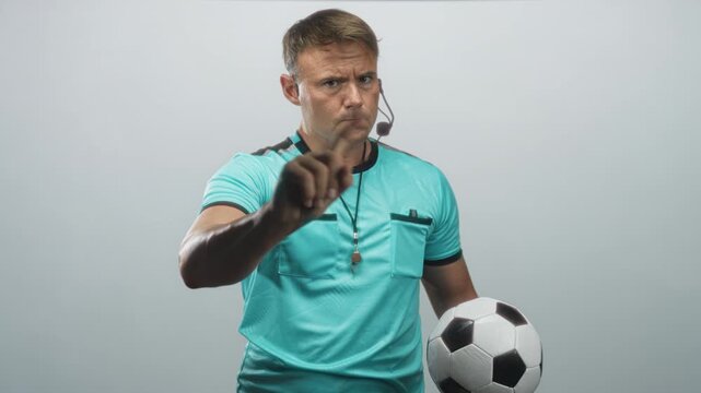 Man referee holding soccer ball, pointing finger and holding whistle near mouth in studio; fairness discipline serious.