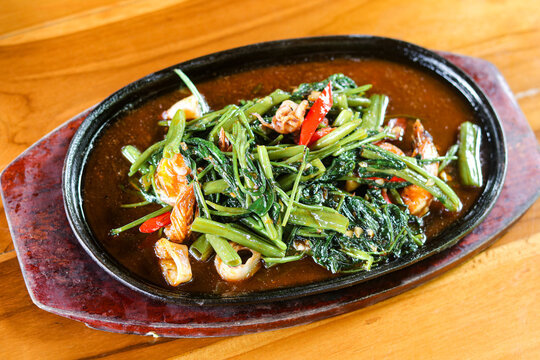 Top view of Kangkung (Water Spinach) Seafood Hotplate, featuring stir-fried water spinach with shrimp, squid rings, and chili, served sizzling on a dark hotplate.