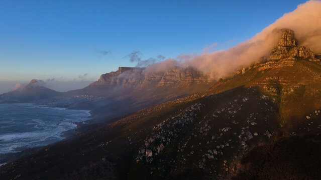 Aerial view of Table Mountain and the Twelve Apostles mountain range with clouds rolling over the peaks during golden hour in Cape Town, Western Cape, South Africa.