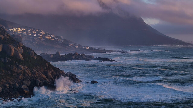 Aerial view of the rocky coastline with crashing waves and houses on a hillside under misty mountains in Cape Town, Western Cape, South Africa.