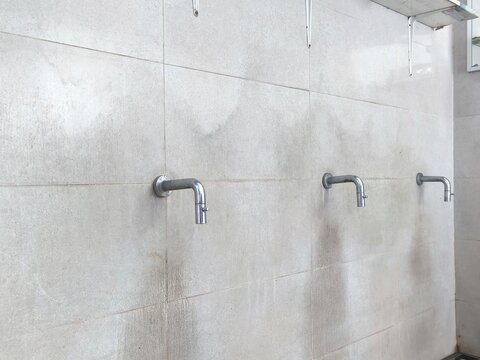 Row of stainless steel water taps installed on a white tiled wall in a public washroom or ablution area.