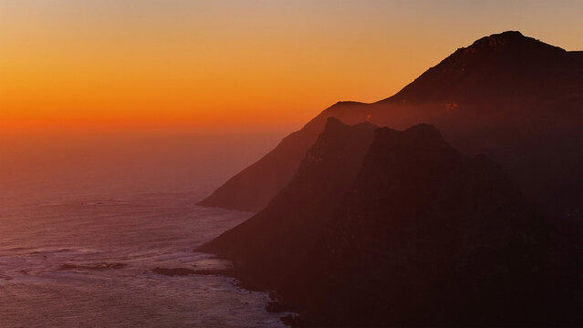 Aerial view of rugged mountain cliffs meeting the Atlantic Ocean under a vibrant orange sunset sky Cape Town, Western Cape, South Africa.