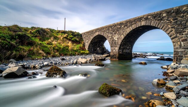 Stone arch bridge spans rocky riverbed with ocean beyond, vibrant sky