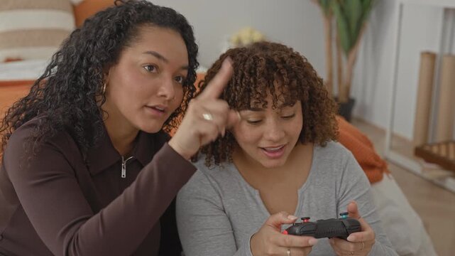 Woman and woman sit on a bed sharing a controller while one points finger to joystick and guides buttons in a bedroom inside building; playful bonding.