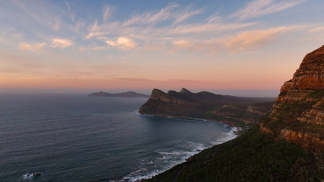 Aerial view of the rugged coastal cliffs and mountains overlooking the Atlantic Ocean under a soft sunset sky in Cape Town, Western Cape, South Africa.