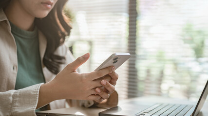 Fototapeta premium Woman using smartphone beside laptop at desk in home office, browsing online, messaging, mobile banking, fintech apps and digital lifestyle communication.