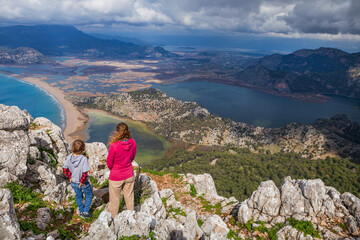 Naklejka premium Young mother and son stand on mountain viewpoint above Dalyan, Turkey, overlooking river delta, lagoon, Iztuzu Turtle beach. Scenic travel moment highlighting family adventure and nature exploration.