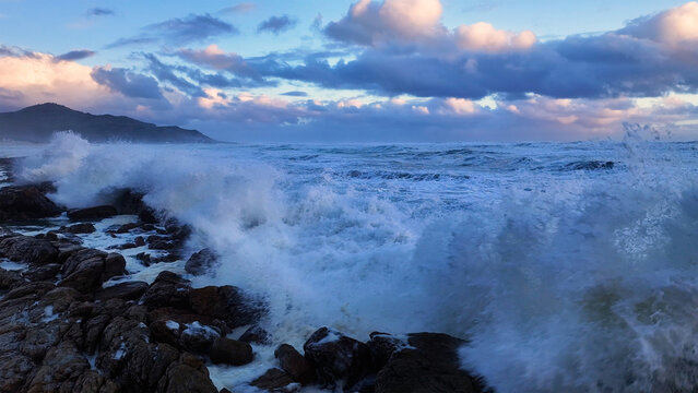 Aerial view of rough ocean waves crashing against a rocky shoreline under a dramatic cloudy sky with distant mountains Cape Town, Western Cape, South Africa.