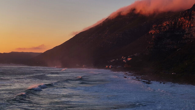 Aerial view of the rugged coastline with waves crashing against the shore and clouds rolling over the mountain peaks during sunset in Cape Town, Western Cape, South Africa.
