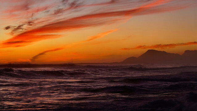 Aerial view of crashing ocean waves under a vibrant sunset sky with orange clouds and mountain silhouettes Cape Town, Western Cape, South Africa.