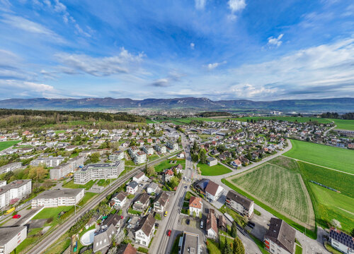 Aerial view of residential houses and lush green fields with the Jura Mountains in the background under a blue sky in Biberist, Solothurn, Switzerland.