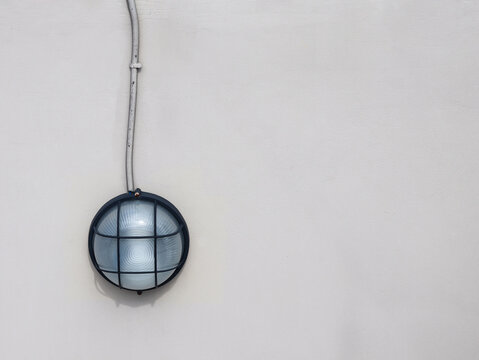 An industrial bulkhead wall lamp mounted on a smooth white wall, features frosted glass enclosed in a protective metal cage, with a visible electrical conduit running vertically above it.