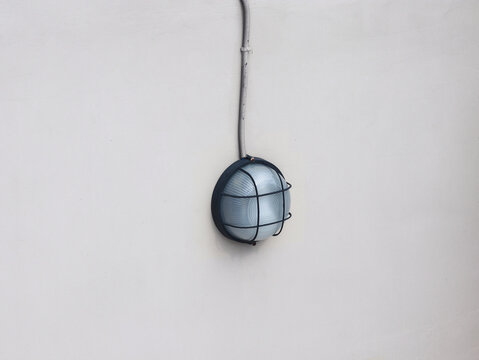Outdoor wall lamp mounted on a clean white wall. The round bulkhead light features frosted glass protected by a dark metal cage, with a visible electrical conduit pipe running vertically above it.
