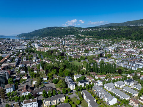 Aerial view of the urban landscape with residential buildings, lush green parks, and rolling hills under a clear blue sky in Biel Bienne, Canton of Bern, Switzerland.