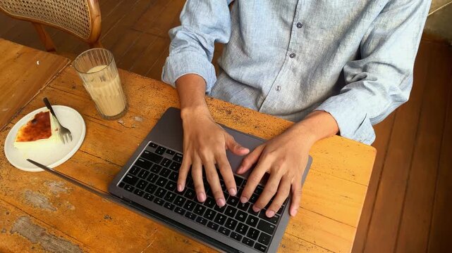 Close up of hands type on laptop keyboard with focused intent. Person wears light blue shirt, seated at wooden table.