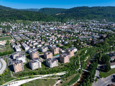 Aerial view of modern residential apartment blocks nestled between a winding river and lush forested hills under a clear blue sky in Biel Bienne, Canton of Bern, Switzerland.