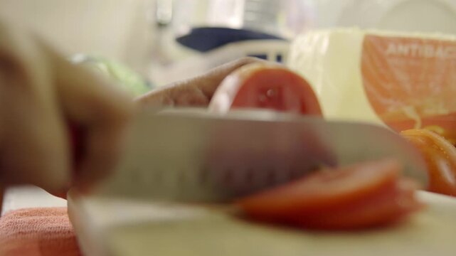 Close view of hand slicing tomato into thin pieces on wooden cutting board, fresh vegetable prepared for cooking or salad, simple food preparation step in kitchen