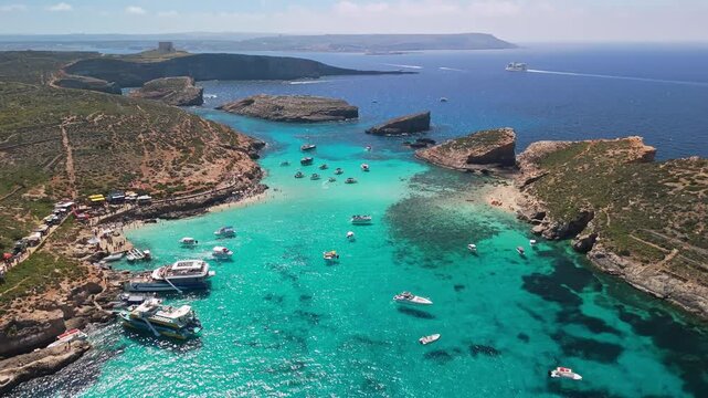 Blue Lagoon, Comino, Malta, features turquoise crystal-clear waters, rocky shores, and anchored boats along the Mediterranean coastline in a picturesque aerial view on a sunny spring day.