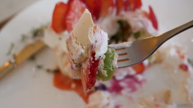 A detailed, slow-motion shot showing a silver fork breaking into a delicate pavlova dessert. The footage captures the contrast between the crispy meringue, soft whipped cream, and vibrant fruit sauces