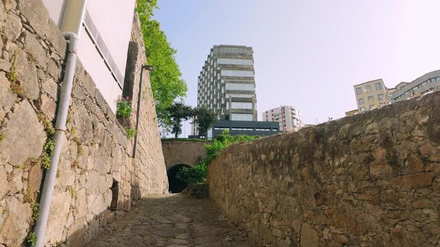 Narrow cobblestone street winding between ancient stone walls on a sunny day in Porto. Distant modern architecture contrasts with historical charm. Green foliage frames the pathway.