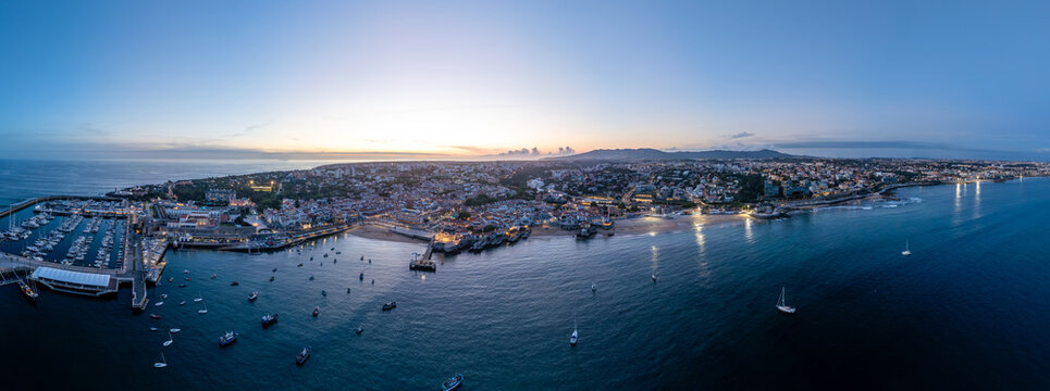 Aerial view of the illuminated coastline, marina with yachts, and sandy beaches at twilight under a clear sky in Cascais, Lisbon, Portugal.