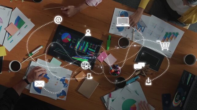 A diverse team engages in a collaborative business meeting, working together over a table filled with charts, graphs, coffee cups, and digital connectivity icons. Raster