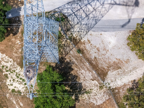 Aerial view of a high-voltage electricity pylon with a stork nest on its arm casting a long shadow over dry terrain and green trees in Palmela, Setubal, Portugal.
