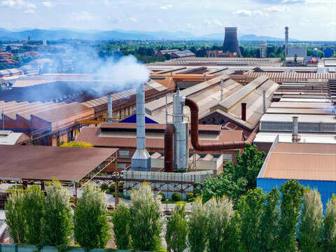 Aerial view of the industrial plant with smoking chimneys, cooling towers, and large warehouses under a blue sky with mountains in the distance in Dalmine, Lombardy, Italy.
