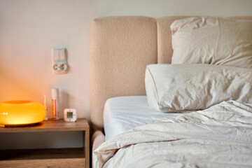 Unmade bed with white bedding, pillows and soft headboard stands beside a wooden nightstand with a warm glowing lamp. Bedroom interior, morning routine, home comfort, sleep and lifestyle concepts © Lazy_Bear