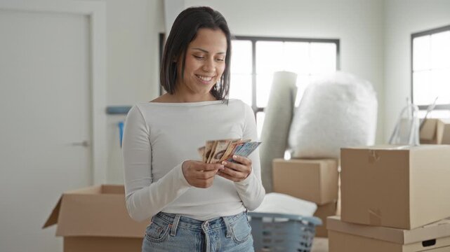 Young hispanic woman counting pesos, hands holding banknotes and fanning bills amid packed moving boxes and laundry basket in a building; relief fresh start.