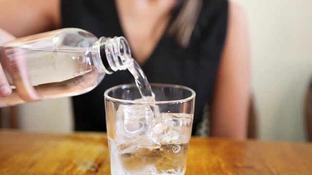 Clear water being poured from plastic bottle into glass full of ice cubes on wooden table, close up, slow motion.