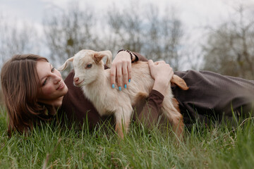 Naklejka premium Young woman lying in grass outdoors, gently embracing a playful goat kid, capturing a warm moment of companionship, nature connection, soft light, relaxed mood, casual lifestyle.